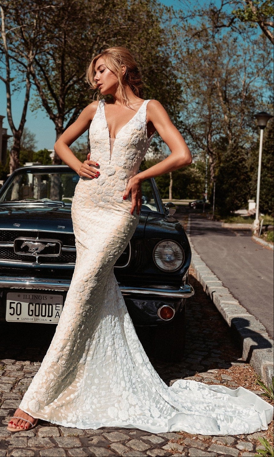 A woman wearing the Long White Mermaid Bridal Gown: Tina Holly BB011 poses before a classic black Ford Mustang on a cobblestone street, framed by trees and charming houses in the background.